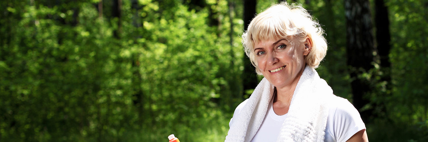 Kvinde der er iført træningstøj og står med en vandflaske i hånden og et håndklæde om nakken